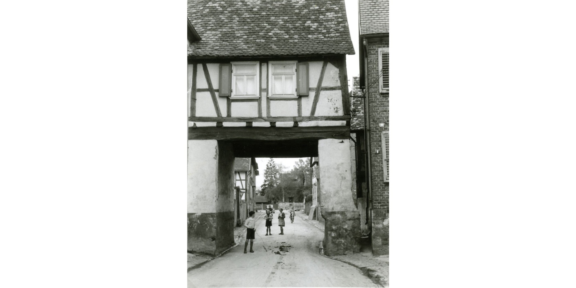 Schwarz-weiß-Aufnahme eines Fachwerkhauses mit einem Durchgang hindurch und spielende Kinder in der Straße