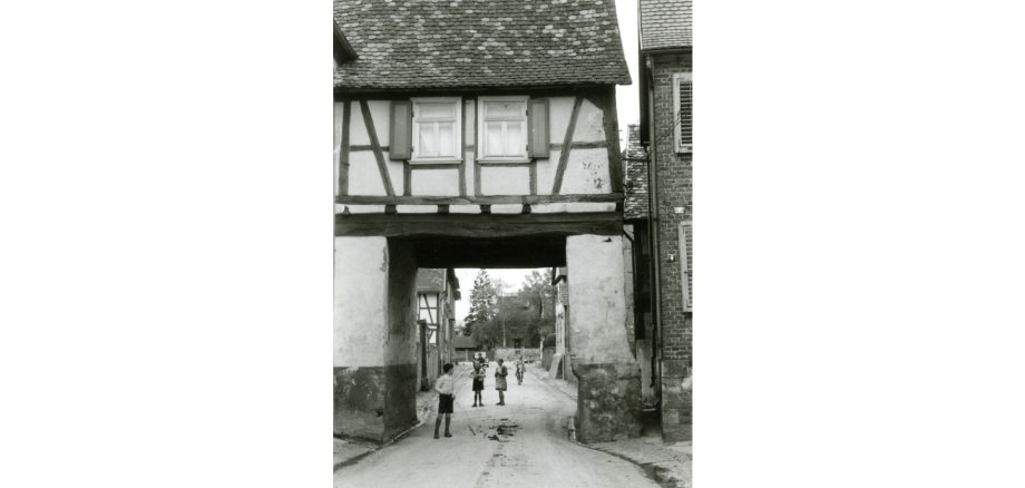 Schwarz-weiß-Aufnahme eines Fachwerkhauses mit einem Durchgang hindurch und spielende Kinder in der Straße